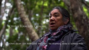 Una mujer de cabello canoso y una bufanda colorida habla en un bosque, representando al pueblo indígena kawésqar. Su nombre, Celina Llanllán Catalán, aparece en pantalla, mientras que los subtítulos en español hablan de ser nómadas del mar.